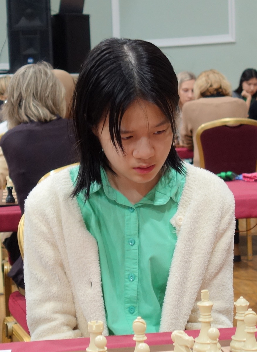 Alice Lee at the board in 2023, looking down at the pieces, in a green shirt and a cream cardigan