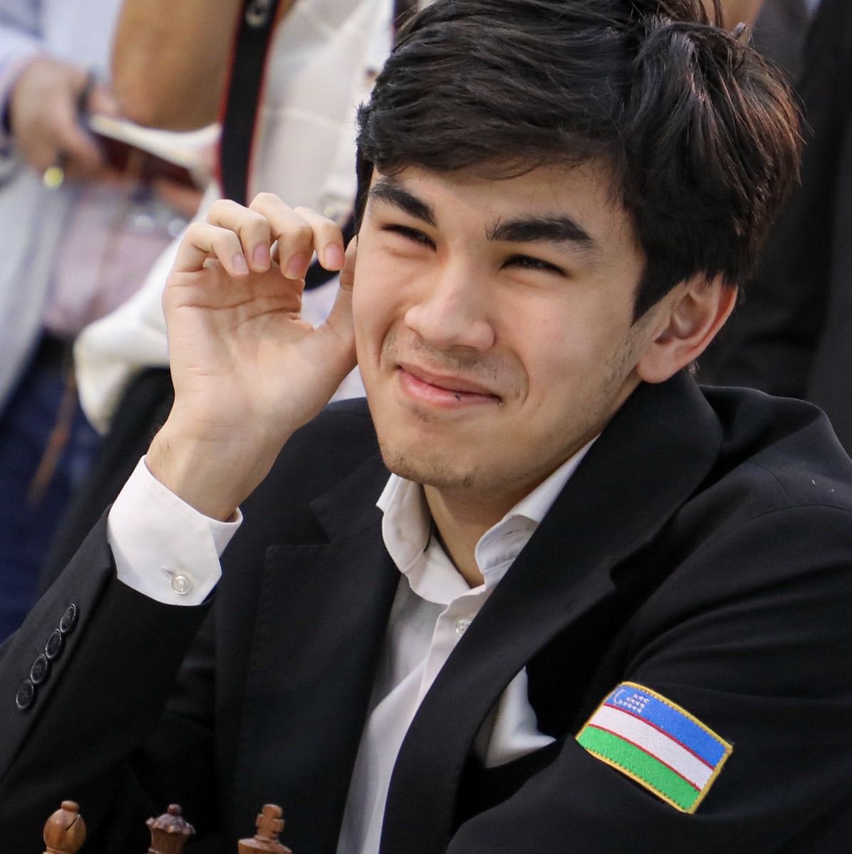 Javokhir Sindarov at the board at the 46th Chess Olympiad in Samarkand, September 2026, smiling, with the Uzbekistan flag on his jacket