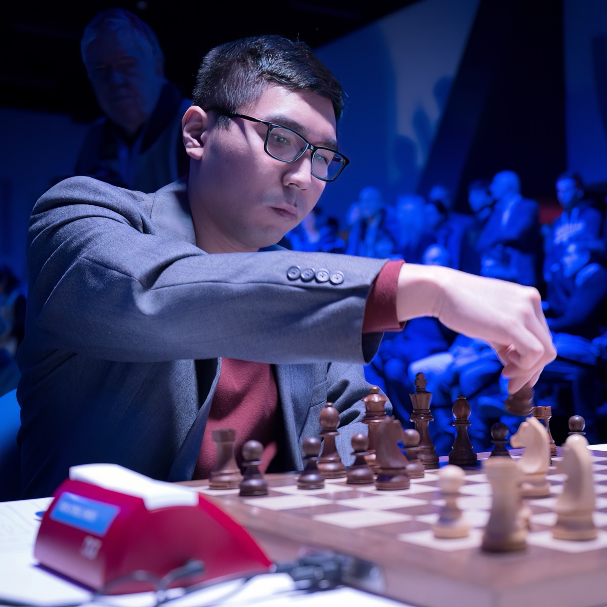 Wesley So reaching to move a piece at the 2023 Tata Steel Chess Tournament, in glasses and a grey jacket